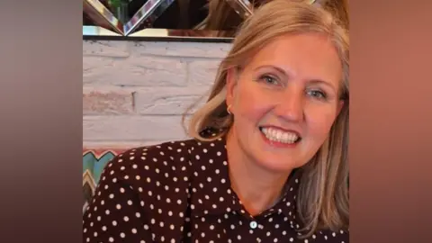 Joanne Mulholland is smiling brightly at the camera. She has long blonde hair and is wearing a brown shirt with polka dots on it. There is a mirrored wall behind her head.