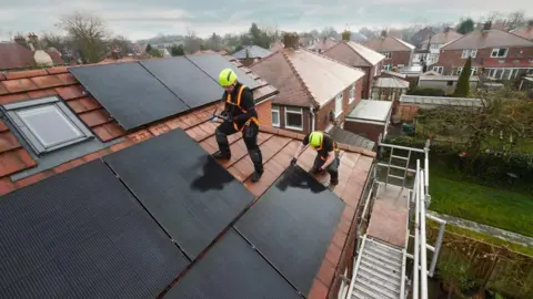 Workmen on the roof of a house installing solar panels while wearing orange harnesses and yellow helmets 