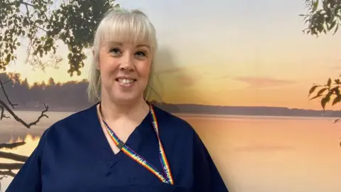 Rebecca Beggan, midwifery programme lead at York St John University stands in front of a mural of a sunset over a lake. She has light hair tied up in a ponytail, and wears navy blue hospital scrubs and a rainbow patterned lanyard.