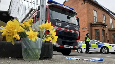 BBC Daffodils in a bucket by the scene of the fire with a fire engine and a policeman also by the cordon