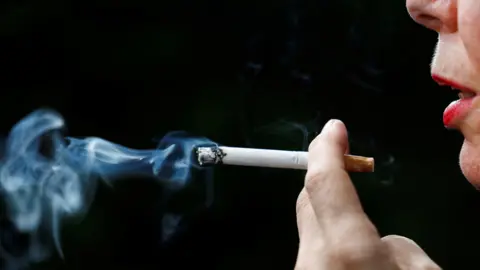 A woman smokes a cigarette in front of a dark background.