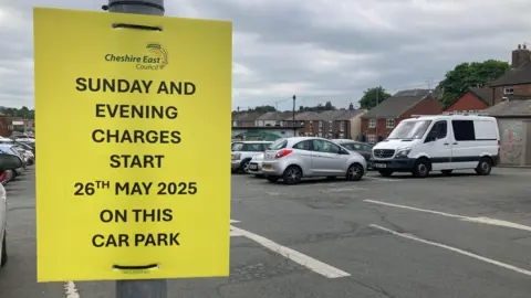 Car park with yellow sign topped Cheshire East Council reading: Sunday and evening charges start 26th May 2025 on this car park