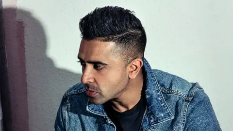 Naroop Jhooti Jay Sean, a man looking to his right, wearing a blue denim top and black tshirt.