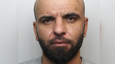 A police close-up image of Khaliz Alshimery.  He has a shaven head, dark brown eyes and dark eyebrows and a dark beard and moustache