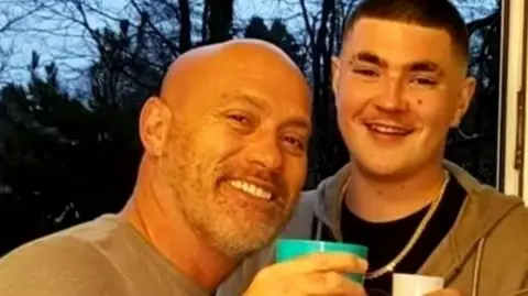 Family A selfie of two men smiling - the one on the left is bald and wearing a grey top while holding a green cup and the one on the right has short black hair, a chain around his neck, a black top and a grey hooded top. He is also holding a white cup. Trees can be seen in the background.