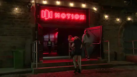 Women walking into the main entrance of Motion nightclub. It is dark and above the main entrance the Motion sign is lit up in red lights.