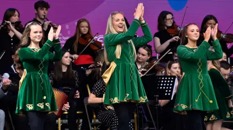 Three girls are pictured on a stage with a seated band behind them. The three are dressed in matching green and gold irish dancing dresses. 