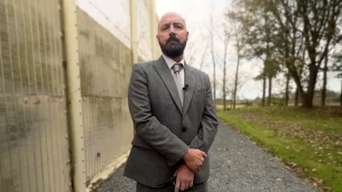 Ben Moore/BBC John De Carteret is wearing a grey suit with a white shirt and pale coloured tie. He has a thick dark beard and is standing in front of a concrete fence