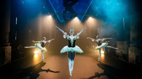 PA Media Three ballerinas in black and white outfits perform dance moves on stage at The Mount Without in Bristol. They are casting shadows due to spotlights and there is dry ice in the air