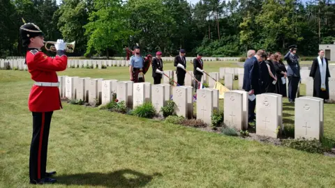 A soldier in a red tunic blowing a horn in front of a row of gravestones with people gathered for a ceremony
