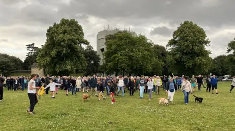 A large group of people including children and dogs walk in a group near the water tower on the downs in Bristol