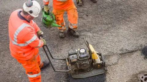 Getty Images A man dressed in orange high-vis and a white hat, uses a hand-held machine to fill in a pothole in a road. 