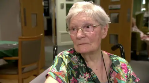 An elderly woman in a floral shirt talks to camera in a lounge