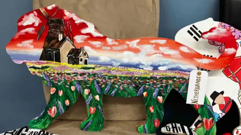A very red, cloudy sky is painted on a lion as well as a wooden windmill, and a field of flowers.