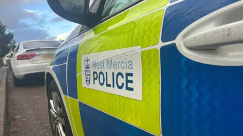 A close-up image of the side of a police car. The vehicle is white with yellow and blue high viz panels on the side. There is also a white West Mercia Police sticker on the side. In front of the police car is a white Audi. 