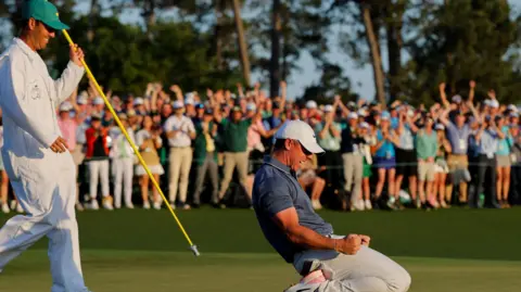 Reuters McIlroy is on his knees with his fists clenched in celebration. He is wearing a white cap, navy polo top and white chino style trousers. He is knelt down on the green and his caddie is behind him wearing a white boiler suit and green cap. He is smiling and holding a yellow pole.