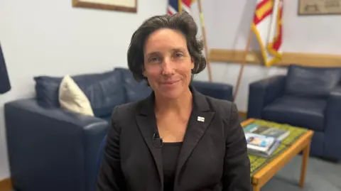 Lindsay de Sausmarez, Policy and Resources President, in a dark blazer sits in an office with blue sofas, a coffee table and flags in the background.
