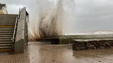 A large wave crashing over a sea wall on to the path. 