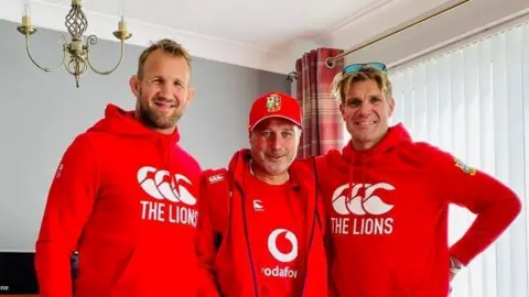 Three men are wearing red Lions rugby clothes. Scott, on the right, is a man in his 40s with blonde hair and sunglasses on his head.