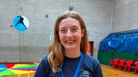The Mae Murray Foundation Woman with blonde hair wearing a blue t-shirt in a gym hall.