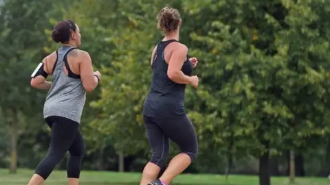PA Media Two women jog through a leafy green park. Both wear vest tops and leggings. One of the women, on the left, also wears a running armband to hold her phone.