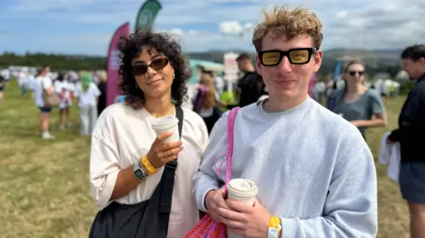 Sarah has dark curly shoulder length hair and is wearing sunglasses, a white t shirt, with a black bag over her shoulder. Dylan, on the right, is wearing yellow lensed sunglasses, has short blonde hair, wearing a grey sweatshirt. They're standing in a crowded field.