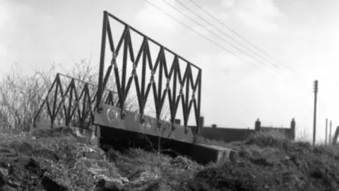 A black and white image of a small bridge with high-sided metal railings on either side and a grass hill