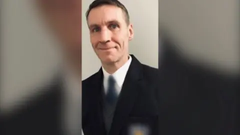 PSNI A man in a white shirt and dark-coloured blazer and tie with short cropped dark hair with grey flecks smiles towards the camera.