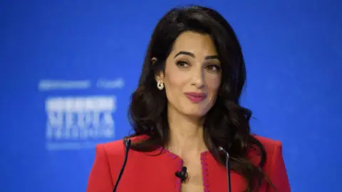 Amal Clooney wears a smart red jacket, and stands at a podium, possibly giving a speech. She has long dark hair which comes past her shoulders.