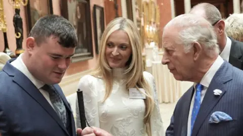 Brandon Hulcoop, who wears a blue suit and holds a stick is seen with Fearne Cotton and HRH King Charles at Buckingham Palace