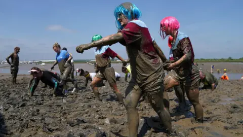 PA Media People walking and crawling through the thick mud during a previous race. In the foreground, two people wearing colorful wigs march through the mud while holding hands.
