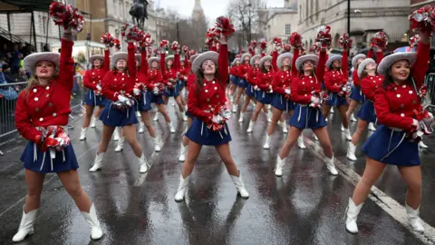 Reuters Three rows of female cheerleaders wearing white boots, blue skirts, red blouses with silver buttons and white Stetson hats, hold aloft their white pompoms. BIg Ben is visible in the background. 