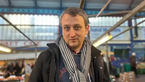 A man with a black and white gingham scarf stands in front of market stalls.