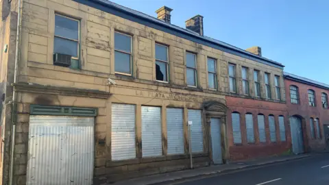 BBC A former manufacturing building is a mix of brown stone and red brick. The windows and gateway on the ground floor have metal shutters covering them. Some of the windows on the first floor are broken. The words La Plata Works are carved into the stone work at the front of the building