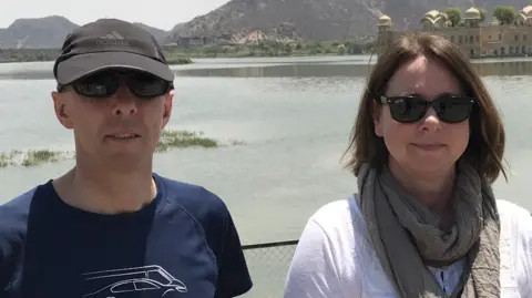 Carol Try A man and woman stand in front of a body of water. The man - Chris - is in a blue Hendy T-shirt, a black cap and sunglasses. The woman - Carol - is wearing a white shirt, a khaki green scarf and sunglasses. She has chin-length brown hair. 