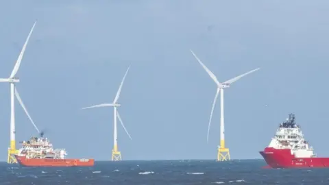 Sea with two red and white boats and large white wind turbines on yellow bases towering above.