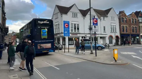 Congestion charge signs at St Clements Street near The Plain Roundabout. Some people are walking on the pavement. A bus is seen travelling up St Clements. A car is driving in the opposite direction. It is a cloudy day.