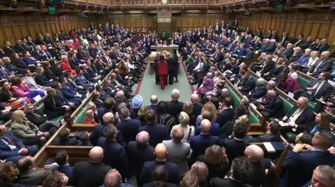 The chamber of the House of Commons in Westminster, London, crowded with Members of Parliament seated and standing around a central aisle. They are gathered to hear the result of the vote on the Terminally Ill Adults (End of Life) Bill. Green benches line both sides, and the Speaker's chair is visible at the far end beneath a clock