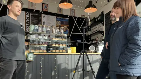 Three people inside a coffee shop, with the owner on the left being filmed. In the background are trays of cakes on the counter top.