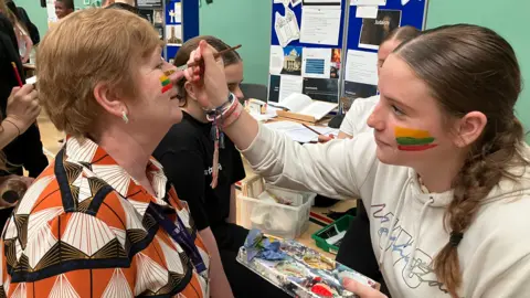 Shariqua Ahmed /BBC A girl painting a woman's face with Lithuanian flag colours - yellow, green and red 