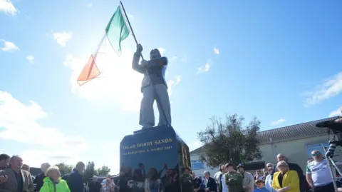 PA Media A low angle photograph of the Bobby Sands statue which holds an Irish tricolour. There are people gathered around at the base of the monument