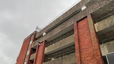 A view looking up at a grey concrete and red brick multi-storey car park with some metal railings. Under a grey sky
