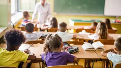 Getty Images Several young children sit in a classroom and face the teacher. They are wearing yellow, purple and white T-shirts and have books and pens in front of them. There are three rows of pupils in the image.