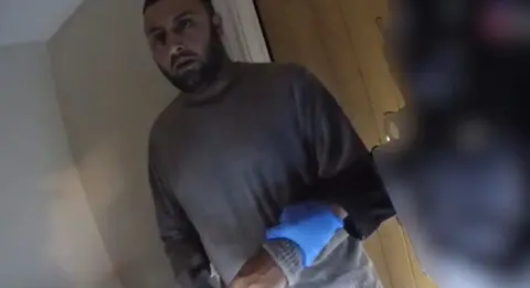 Leicestershire Police A man in grey jumper being arrested inside his house by an officer in a blue latex glove.