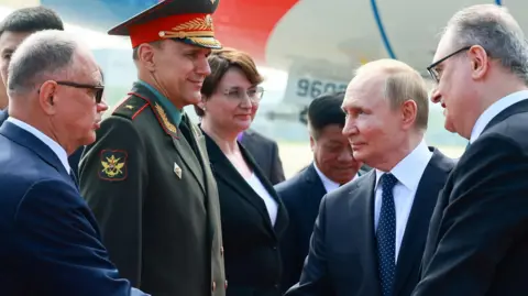 Reuters Russian leader Vladimir Putin shakes hands with officials during a welcoming ceremony. A man wearing green military uniform and hat looks at him from the side. Putin is wearing a dark suit, with a dark tie and light blue shirt.