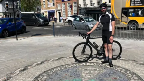 Tom Jackson/BBC Richard Slade is standing in a town centre in front of a mosaic on the pavement.
He is holding a black bicycle. He is wearing a tight-fitting blue and white cycling top and shorts, a bicycle helmet and has sunglasses on. He has a greying beard and moustache.