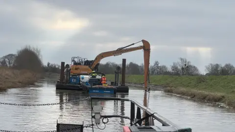 In the centre of the photograph is a digger floating on the river, with a long arm scraping the banks clear. The photo has been taken from a boat behind. Either side is the green river bank and a number of trees.