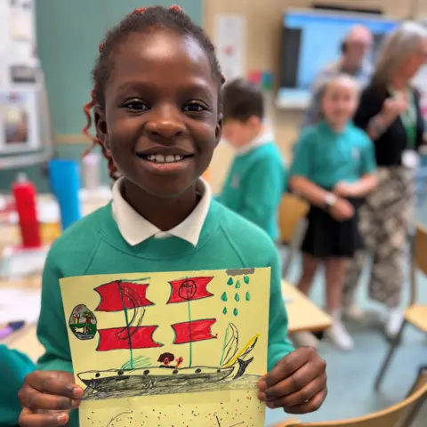 Girl in school uniform smiling at camera and holding a drawing of a Tall Ship.