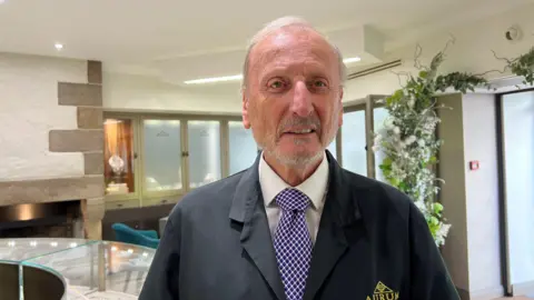 BBC Richard Blampied is looking directly at the camera, wearing a black coat bearing the Aurum logo. He has a purple tie and a white shirt. He is standing inside.