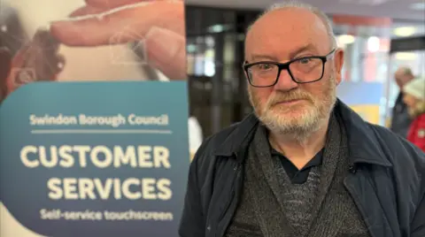 A man wearing glasses and a grey jumper stands next to a sign which says "Customer Services". He has a medium-length beard which is light brown and grey.